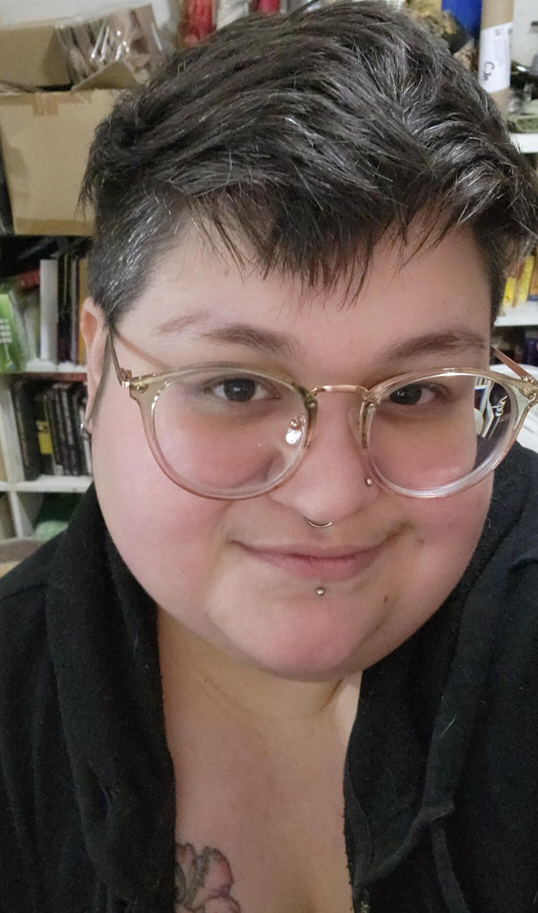 A light skinned Latine with brown hair, large glasses, a nose piercing, a silver septum ring, and lower lip stud.